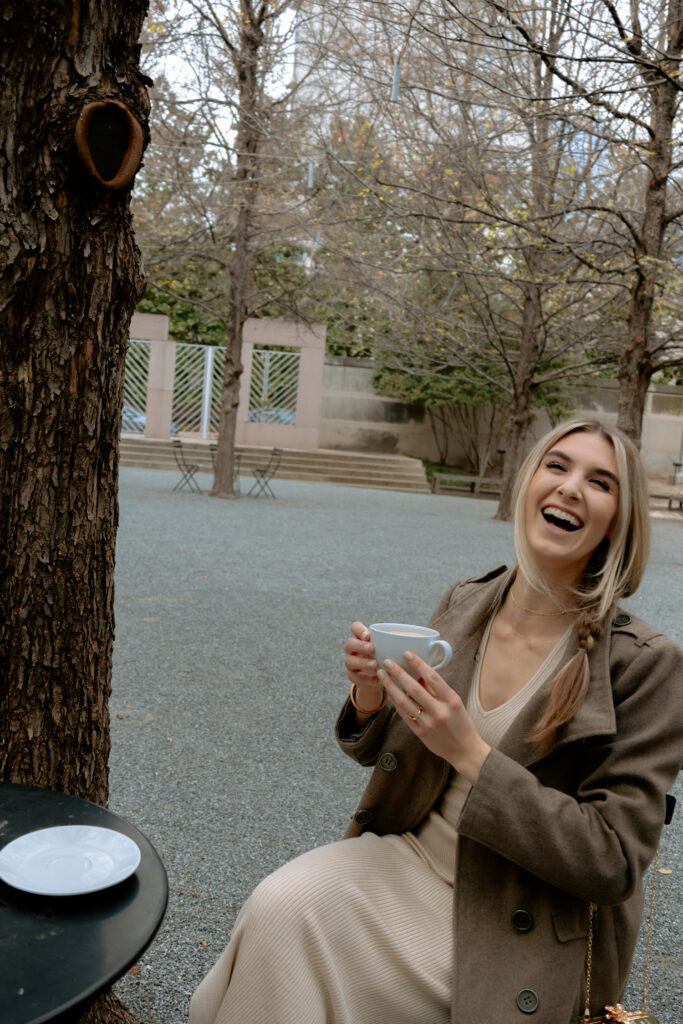 Photo of woman sitting, holding a coffee cup laughing at the camera