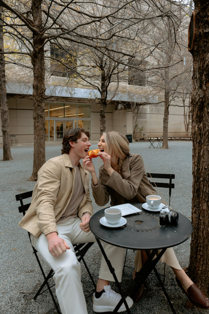 Photo of engaged couple sitting at a table together both biting into a croissant