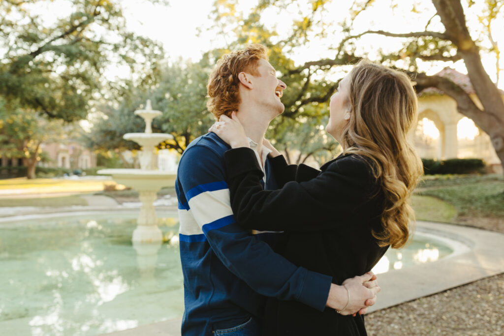 Man and woman with arms wrapped around each other laughing in front of a pond as they just got engaged.